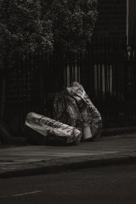 A black and white photograph showing two large, crumpled rubbish bags leaning against a wooden fence on a paved sidewalk. The bags appear to be made of thick plastic material, with some branding or text partially visible on the surface. The surrounding environment includes a tree with dense foliage in the background, and an adjacent building with dark window frames partially visible behind the fence. The scene is outdoors, likely in a residential or urban area, with the rubbish bags indicating an instance of waste accumulation that may require collection by local or private rubbish removal services such as Rubbish Clearance Harringay, especially where access issues could complicate waste disposal.