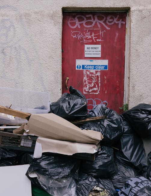 A pile of black garbage bags and assorted waste materials, including cardboard and foam, are stacked in front of a red metal door set into a textured, off-white wall exterior. The bags vary in size and are partly overlapping, with some leaning against the door. The door features graffiti tags, a white sign indicating 'No Money' and warning about fines, as well as a blue 'Keep Clear' sticker. The surrounding environment suggests an alleyway or service entrance area, typical for private waste collection or rubbish clearance services, with the waste likely awaiting removal by a professional rubbish clearance company such as Rubbish Clearance Harringay. The scene conveys a typical example of rubbish awaiting disposal, highlighting on-site waste accumulation that may require alternative waste handling solutions for efficient clearance and environmental management.
