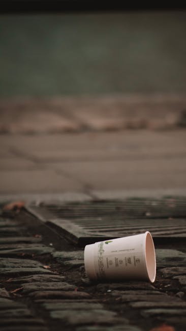 A black and white photograph showing two large, crumpled rubbish bags leaning against a wooden fence on a paved sidewalk. The bags appear to be made of thick plastic material, with some branding or text partially visible on the surface. The surrounding environment includes a tree with dense foliage in the background, and an adjacent building with dark window frames partially visible behind the fence. The scene is outdoors, likely in a residential or urban area, with the rubbish bags indicating an instance of waste accumulation that may require collection by local or private rubbish removal services such as Rubbish Clearance Harringay, especially where access issues could complicate waste disposal.
