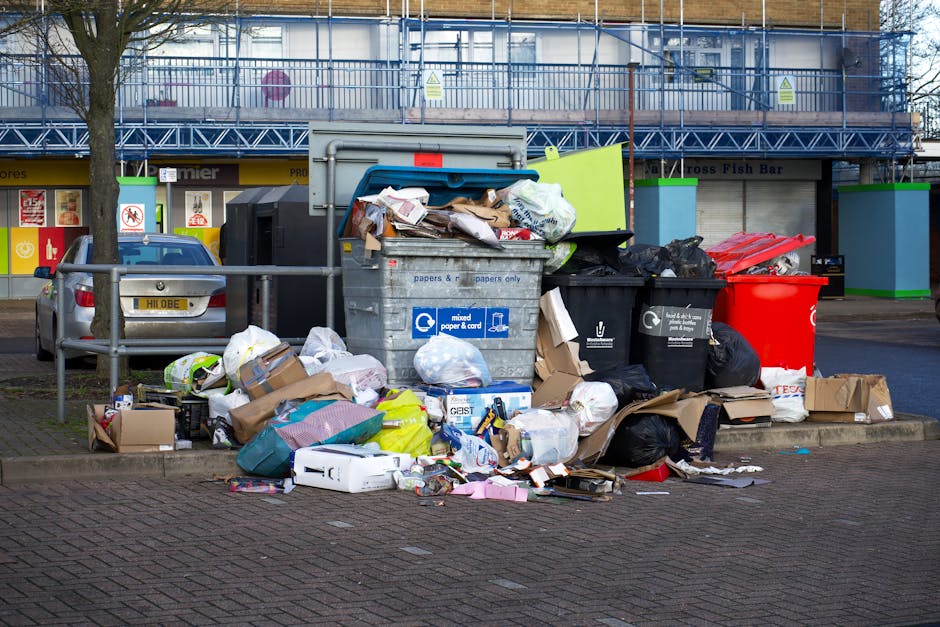 A collection of overflowing rubbish bins and scattered waste surrounds a designated recycling area in an urban setting. The waste includes black, grey, and red plastic bins, with some lids open and contents spilling out. The black and grey bins are labeled for mixed paper and cardboard, while the black bin is for other general waste. Various types of waste such as flattened cardboard boxes, paper bags, plastic packaging, and scattered loose papers are piled around the bins on a paved sidewalk section. Additional waste items, including plastic containers, paper waste, and a few small boxes, are strewn across the ground, indicating significant buildup. Behind the rubbish, there is a metal railing separating the waste area from a parking lot with a parked silver car. In the background, there is a row of commercial units with closed shutters, and above them, a building with scaffolding and construction barriers painted blue and green. The overall scene suggests an example of improperly managed waste collection, highlighting the need for alternative waste handling or private rubbish removal services in the area.