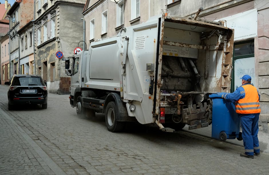 A sanitation worker dressed in a high-visibility orange safety vest and blue coveralls is shown standing on a narrow cobbled street, tipping a large blue recycling bin into the opening of a white waste collection truck. The truck is equipped with a rear-loading mechanism and shows signs of rust and wear inside the hopper. The scene is set in an urban environment with old, multi-story buildings lining the street, some with faded, peeling paint and exposed brickwork. A black passenger car is parked on the street beside the truck, and street signs, including a no parking sign, are mounted on the buildings. Natural daylight illuminates the scene, highlighting the textures of the cobblestones, the rough surface of the buildings, and the metallic surface of the waste machinery, illustrating an instance of independent refuse collection that supports alternative waste handling methods outside of municipal sanitation services, related to rubbish removal services typical in the Harringay area.