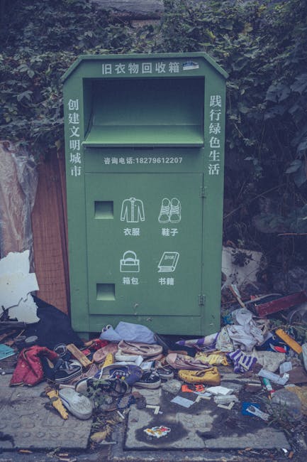 The image depicts a green old clothing recycling box positioned outdoors on a paved surface, with a dense background of greenery and foliage behind it. The box features Chinese characters on the top and along the sides, indicating it is used for collecting discarded clothing and accessories. The front panel has separate slots for different items, including a slot labeled for clothes, shoes, bags, and books, each accompanied by relevant simple line icons. In front of the box, there is a significant amount of mixed rubbish scattered across the pavement, including various discarded shoes, clothing, plastic containers, and paper items, some of which appear to be strewn haphazardly or in disorganized piles. The scene suggests an instance of improper waste disposal related to clothing and miscellaneous rubbish, highlighting issues associated with private waste handling and loose rubbish collection, with Rubbish Clearance Harringay occasionally dealing with such scenes in their rubbish removal services. The overall lighting is natural, with soft daylight illuminating the area, emphasizing the contrast between the neatly designed recycling box and the surrounding clutter.