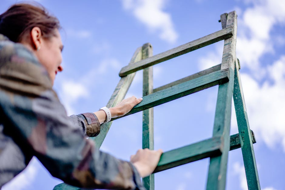 A person with brown hair, wearing a multi-colored jacket and a white wristwatch, is seen climbing a weathered wooden ladder outdoors. The ladder has greenish-blue paint that is chipped and faded, revealing the natural wood underneath. The individual is grasping the ladder’s side rails with both hands, with their left hand visible and positioned near the top, while their face is partially turned away. The background features a bright blue sky with scattered white clouds, indicating a clear day. The environment appears to be an open outdoor space, possibly a garden or backyard, with no other structures or objects immediately visible. The scene's lighting suggests daytime, with natural sunlight illuminating the ladder and the person. This visual aligns with themes of DIY project, site clearance, or on-site rubbish removal, often associated with private waste handling and independent collection services that companies like Rubbish Clearance Harringay may facilitate through their rubbish removal services.