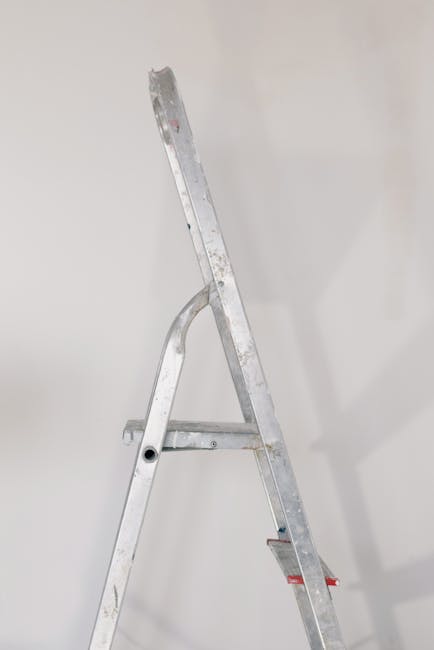 A close-up view of a silver metal ladder with visible scratches and signs of use, positioned upright against a plain, light-colored wall. The ladder has a primary vertical section with a textured, slightly worn surface, and horizontal steps with a matte finish. The side rails are connected with metal brackets, and a curved support connects the two vertical sides approximately halfway up. The feet of the ladder are not visible in the image. The environment appears to be indoors with soft, diffuse lighting that creates subtle shadows on the wall behind the ladder. This scene is typical of a setting where a ladder might be used for maintenance, repairs, or cleaning, which could relate to on-site rubbish clearance or work involving the removal of household or building waste, as seen in professional rubbish removal services such as those provided by Rubbish Clearance Harringay.
