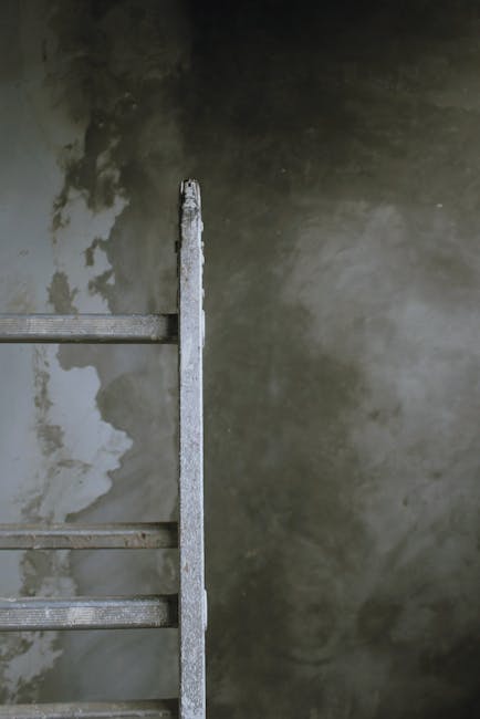 A person with brown hair, wearing a multi-colored jacket and a white wristwatch, is seen climbing a weathered wooden ladder outdoors. The ladder has greenish-blue paint that is chipped and faded, revealing the natural wood underneath. The individual is grasping the ladder’s side rails with both hands, with their left hand visible and positioned near the top, while their face is partially turned away. The background features a bright blue sky with scattered white clouds, indicating a clear day. The environment appears to be an open outdoor space, possibly a garden or backyard, with no other structures or objects immediately visible. The scene's lighting suggests daytime, with natural sunlight illuminating the ladder and the person. This visual aligns with themes of DIY project, site clearance, or on-site rubbish removal, often associated with private waste handling and independent collection services that companies like Rubbish Clearance Harringay may facilitate through their rubbish removal services.