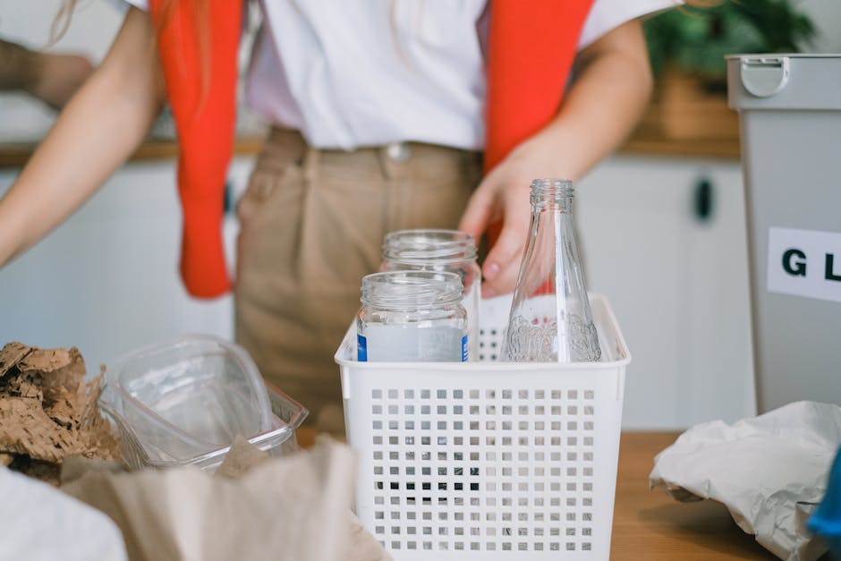 A person in casual clothing, wearing a white shirt and beige trousers, stands behind a cluttered workspace with various glass jars and containers. The jars are transparent and appear to be empty or partially filled, with some having metal lids and others without. The jars are placed in a white plastic basket with a grid pattern, positioned centrally in the image. To the left, there are additional items such as crumpled paper and irregularly shaped pieces of cardboard or natural material, indicating waste or recyclables. On the right side, part of a gray recycling bin with a label is visible, subtly suggesting waste management or collection actions. The background shows a wooden surface and a slightly blurred indoor environment, with lighting that appears natural and even. This scene visually relates to waste sorting, recycling, or household rubbish collection, aligning with independent or alternative waste handling services such as those offered by Rubbish Clearance Harringay, within a domestic or small-scale context.