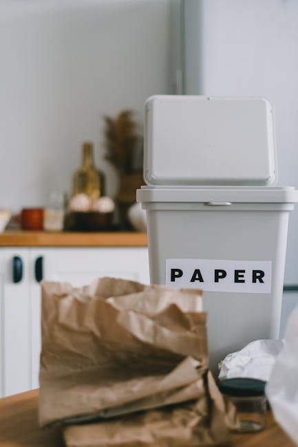 A white plastic recycling bin with a hinged lid, positioned on a wooden surface indoors, features a label reading 'PAPER' in black capital letters. In the foreground, there is a crumpled brown paper bag with some waste paper and small miscellaneous items placed nearby. The background shows a kitchen setting with a white kitchen unit, some blurred decorative objects, and a wooden countertop. The scene depicts an organizational setup for separating household paper waste, which relates to waste management practices such as on-site clearance or alternative disposal methods handled by services like Rubbish Clearance Harringay. Natural lighting illuminates the scene softly, emphasizing the textures of the paper bag and the smooth surface of the recycling bin. The overall environment suggests an indoor space prepared for rubbish collection, highlighting typical household waste separation activities consistent with local or private waste handling regulations.