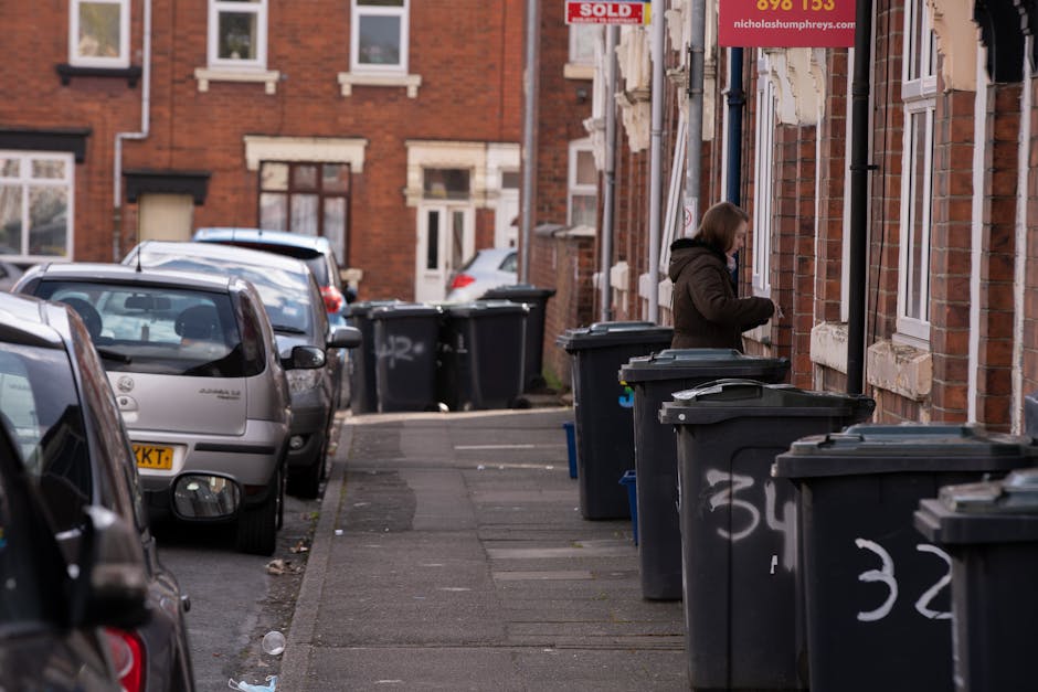 A row of black wheelie bins lined up along the sidewalk outside a brick residential building, with some bins partially open or closed and graffiti markings visible on their sides. A woman dressed in a dark coat is standing next to one bin, appearing to be opening or placing rubbish inside. The background shows parked cars along the curb, with a mix of older and newer vehicles, and additional houses made of red brick with white window frames and decorative stone detailing. The pavement and street surface are visible, with some litter scattered on the ground. The scene is illuminated by natural daylight, indicating daytime, and the environment suggests an urban residential area where private waste collection services, such as those offered by Rubbish Clearance Harringay, are used for rubbish disposal rather than official municipal collections.