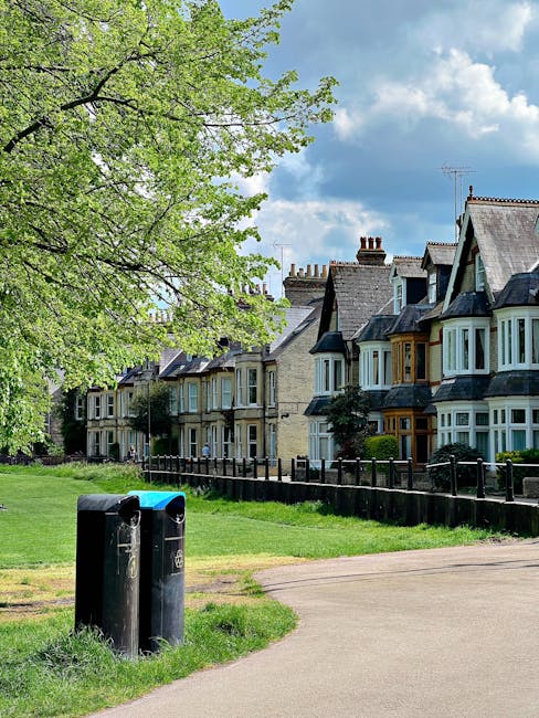 A row of traditional terraced houses with bay windows and pitched slate roofs is visible in the background, situated along a residential street. In the foreground, there is a green area with a large leafy tree on the left side, providing shade and greenery. Two black communal rubbish bins with slightly weathered surfaces are positioned on a grassy patch near a paved pathway that curves gently to the right. The rubbish bins are close to the pathway's edge and located on the grass, adjacent to a low wooden fence that separates the green space from the front gardens of the houses. The sky above is partly cloudy with patches of blue, and the lighting suggests a bright, clear day. The scene is a typical suburban area, with a quiet, well-maintained environment suited for residents and waste collection services such as those provided by Rubbish Clearance Harringay, supporting alternative waste handling outside of municipal rubbish collection.