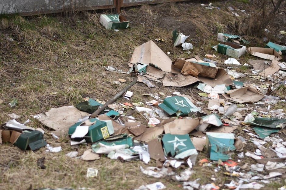 An outdoors scene showing a patch of uneven ground with scattered debris, including crushed and torn cardboard boxes primarily green with white star logos, small pieces of paper, and bits of broken packaging material. Some boxes are partially flattened or ripped open, revealing their contents. The scene includes a mix of brown, beige, and white paper scraps, with a few transparent plastic bottles and small miscellaneous items mixed in. In the background, a section of a metal fence or barrier is visible at the top edge, while sparse grass and bare soil cover the ground surrounding the rubbish. The lighting appears natural, likely daylight, highlighting the disorganized and neglected state of the area, which could be a site needing rubbish clearance or private waste disposal services, as offered by companies like Rubbish Clearance Harringay.