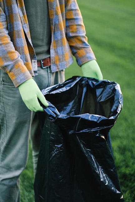 Close-up of a person wearing a yellow and grey checkered shirt, with a green jacket underneath, and grey trousers secured with a brown belt, standing outdoors on green grass. The individual is holding open a large black rubbish bag lined with a shiny black inner layer, with both hands wearing light green gloves made of rubber or similar material. The rubbish bag is empty and prepared for waste collection. The background features a blurred expanse of well-maintained grass, indicating an outdoor setting likely in a garden or park context. The scene suggests an activity related to waste disposal or garden clearance, corresponding with private rubbish removal services, emphasizing the safe handling of waste materials. Visible lighting appears natural, suggesting daylight conditions, and the overall environment appears clean and orderly, suitable for environmentally responsible rubbish clearance.