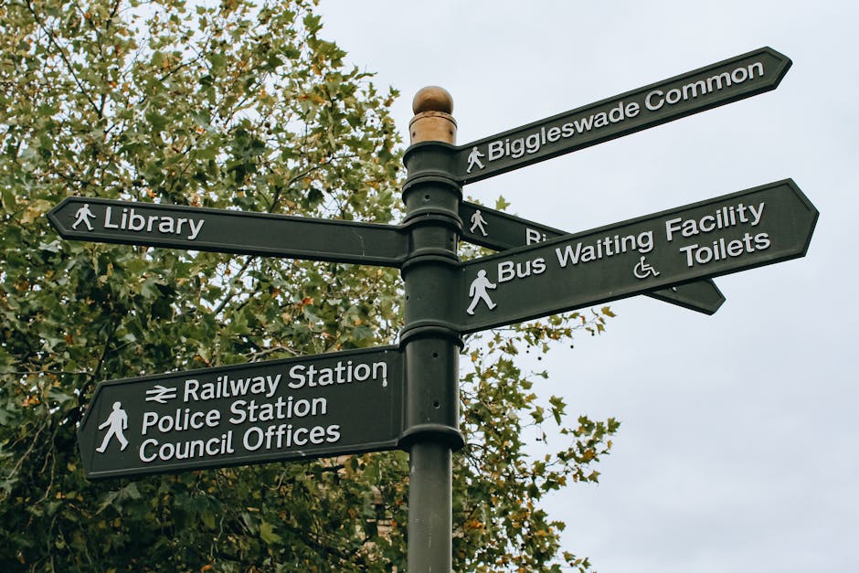 A black metal directional signpost featuring multiple rectangular panels with white text and symbols, positioned outdoors against a background of leafy trees and an overcast sky. The signpost indicates various local destinations, including Biggleswade Common, Library, Railways Station, Police Station, Council Offices, and Bus Waiting Facility with Toilets. Each panel is mounted horizontally on a central pole, which has a rounded finial at the top, and includes pedestrian symbols next to relevant locations, as well as a symbol for wheelchair accessibility on the Toilets panel. The environment appears urban, possibly within a public area or park, and the signpost is clean and well-maintained, consistent with municipal signage commonly used for guiding residents and visitors. This setup could relate to local municipal wayfinding or supplementary signage supporting independent navigation, which aligns with the context of managing or accessing town amenities in a broader urban or suburban setting, including services related to waste management and rubbish collection points.